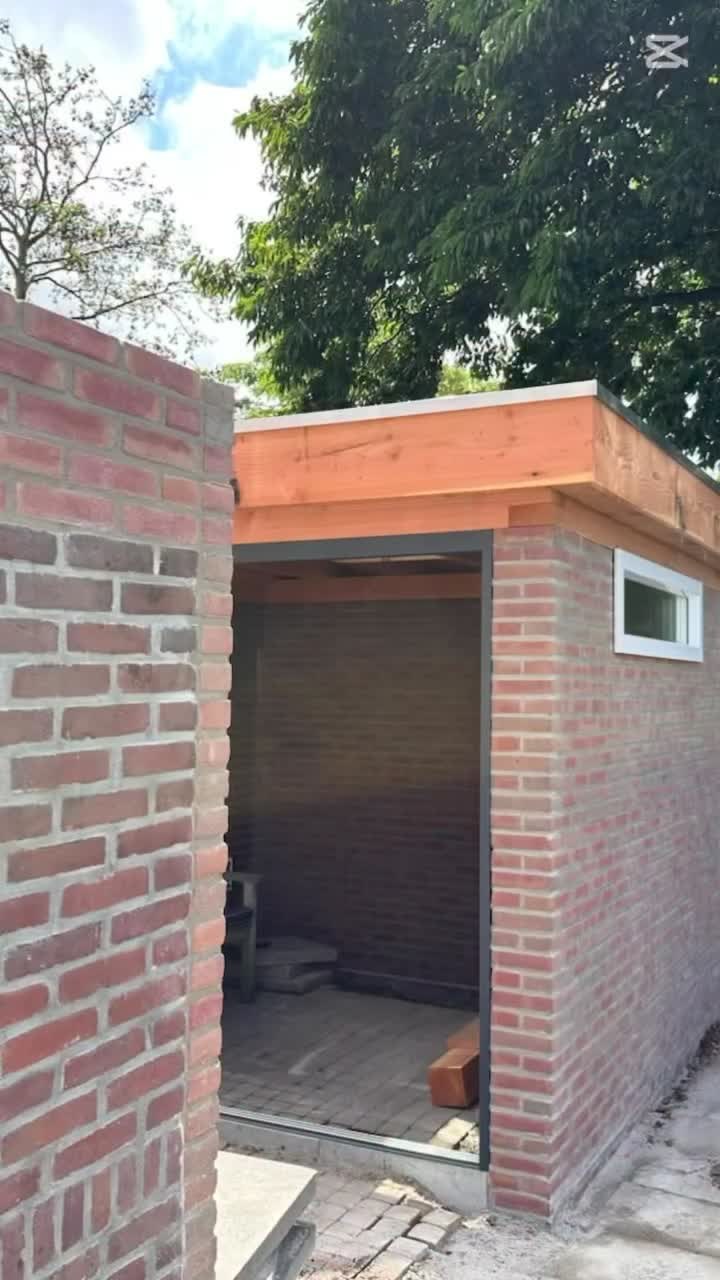 Gemetseld tuinhuis van rode handvormsteen met een plat dak en een open deur, naast een tuinmuur