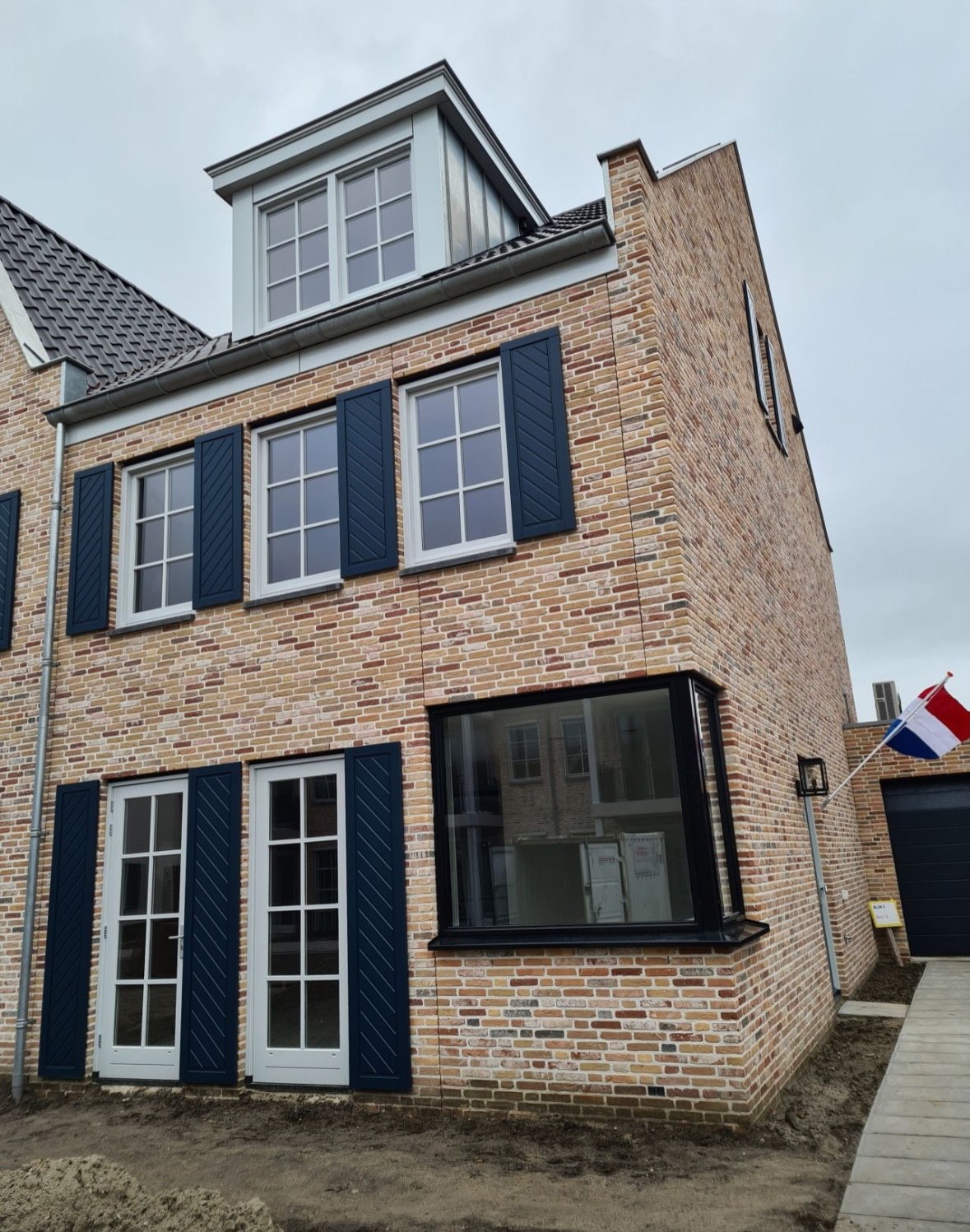 Nieuw gemetselde woning met lichte gevelsteen, donkerblauwe luiken en een erker, met een Nederlandse vlag op de steiger